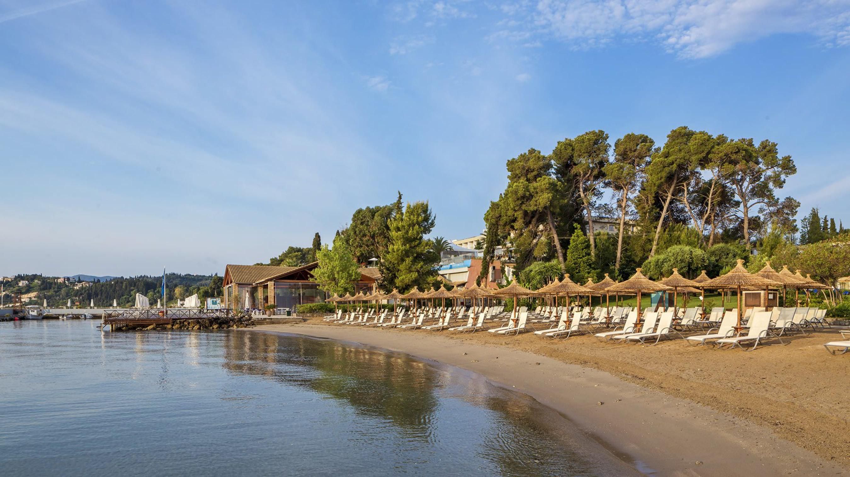 A beach with sun loungers and umbrellas, a building with a sloped roof, and trees in the background.