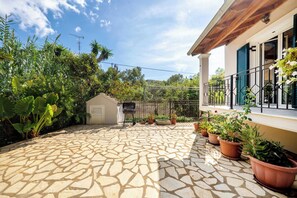 A stone patio with a barbecue, potted plants, and a view of a house with a balcony. - Kallithea House Corfu - Courtyard & Beach Days (Gouvia)