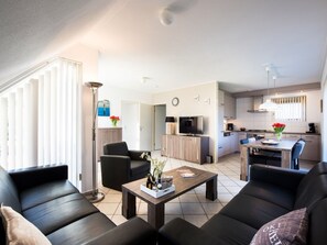 Ein modernes Wohnzimmer mit einer Couch, einem Couchtisch und einem Fernseher. Im Hintergrund ist eine Küche mit Schränken und einem Essbereich zu sehen.