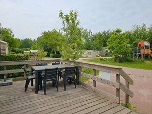 A wooden deck with a table and chairs, surrounded by a wooden fence, with a playground and greenery in the background. - Holiday Home With 1 Bedroom (Oostkapelle)