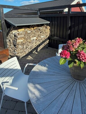 Un patio con una mesa redonda y sillas, una pila de leña y una maceta con flores rosadas.