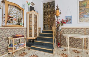 A staircase with ornate metal railings, a wooden cabinet with glass doors, a small table with a vase of flowers, and a patterned wallpaper wall. - Group Accommodation Aquarium | 28 Persons | Sauna | in Monschau (Monschau)
