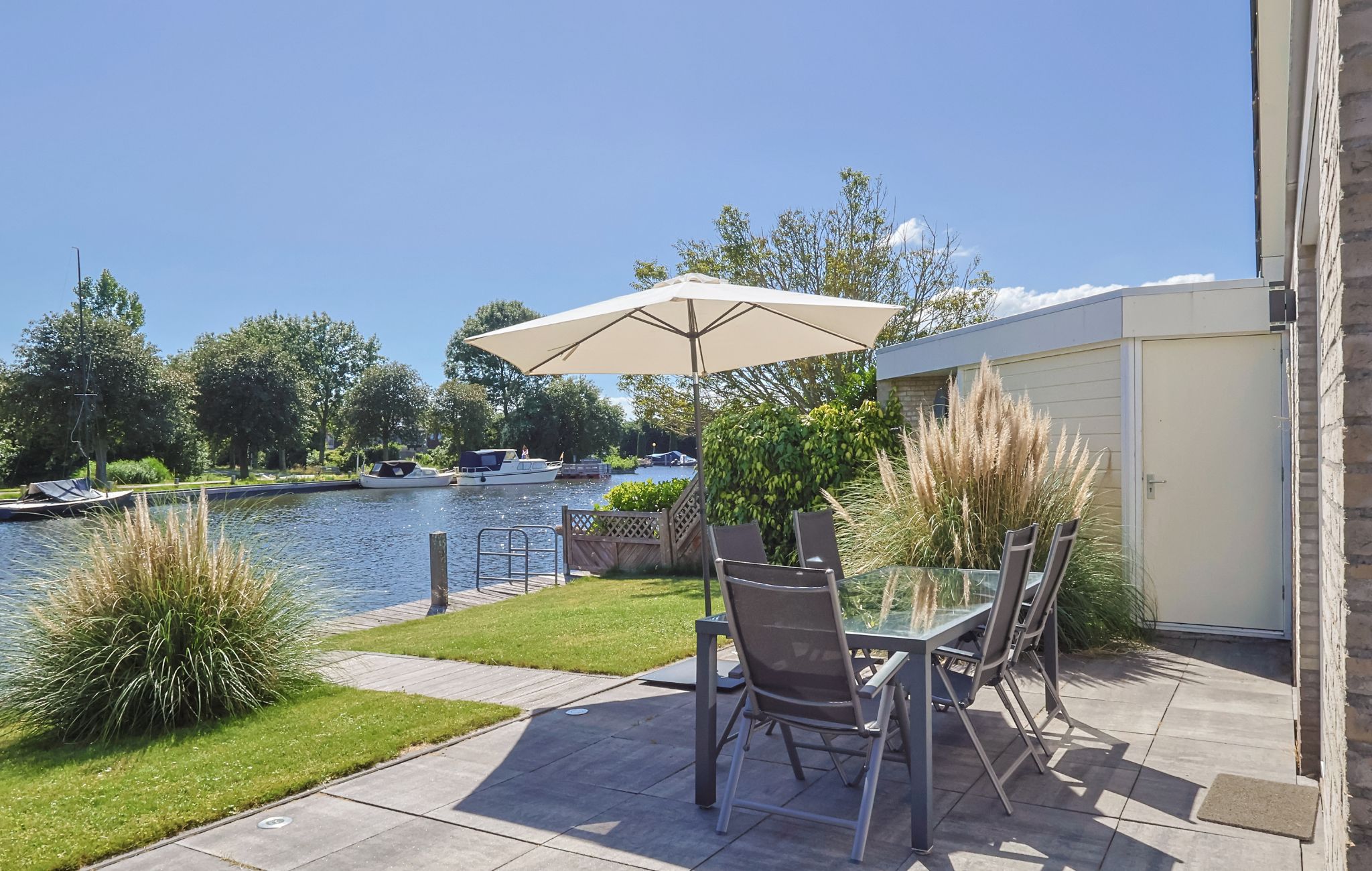 An outdoor patio with a glass table and chairs, a beige umbrella, and a view of a lake with boats.