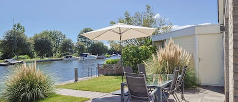 An outdoor patio with a glass table and chairs, a beige umbrella, and a view of a lake with boats.