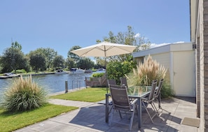 An outdoor patio with a glass table and chairs, a beige umbrella, and a view of a lake with boats. - Watersnip - Enjoying the water (Lemmer)