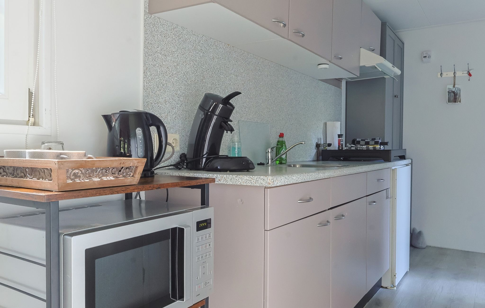 Una cocina con microondas, tetera y cafetera sobre la encimera, un lavabo con grifo y armarios con un salpicadero de aspecto moteado.
