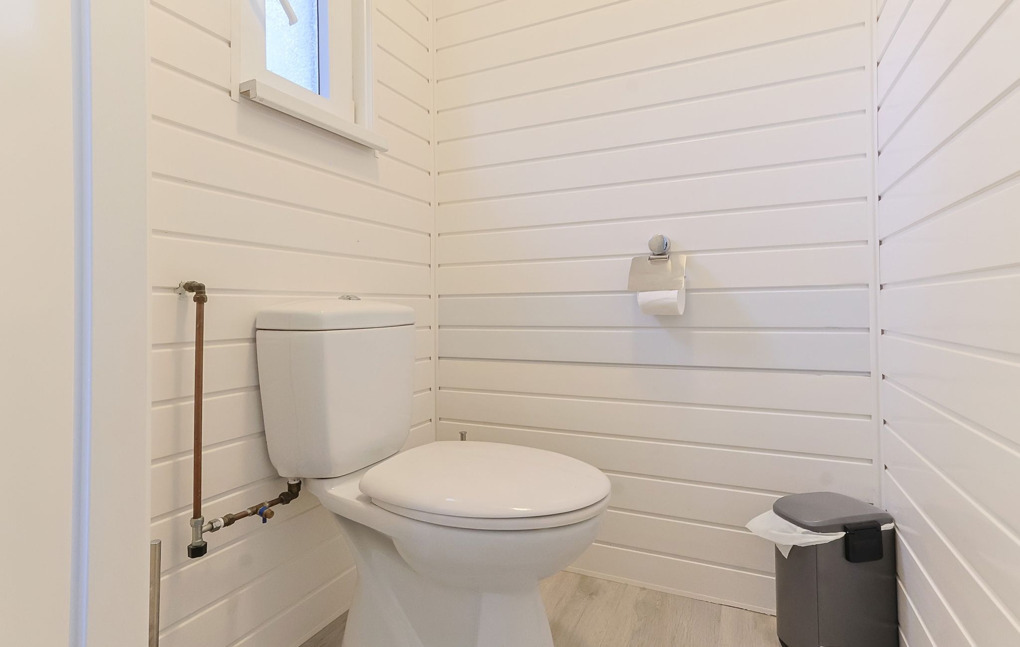 Et lille badeværelse med hvide paneler, toilet, skraldespand og et vindue.