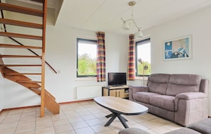 Ein Wohnzimmer mit einer Couch, einem Fernseher, einem Holztisch, einer Treppe und einem Fenster mit Vorhängen.