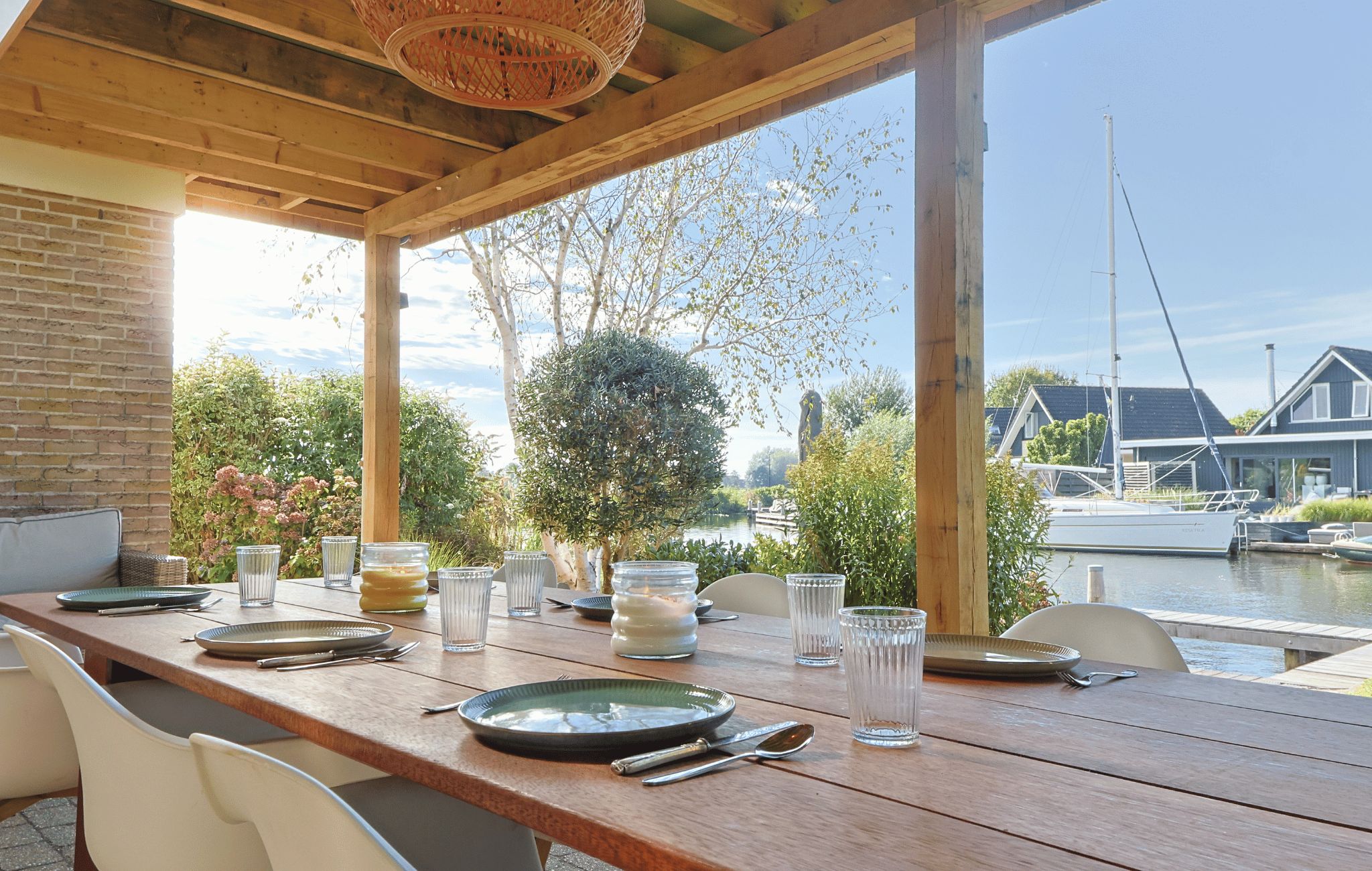 Un área de comedor al aire libre con una mesa de madera dispuesta para dos personas, con vista a un canal con barcos y casas al fondo.