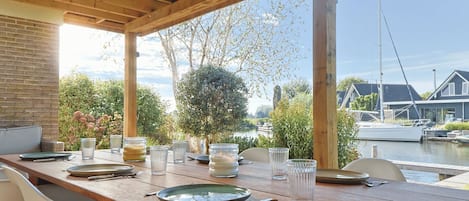 An outdoor dining area with a wooden table set for two, overlooking a canal with boats and houses in the background.