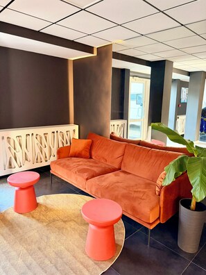 A modern lobby with a sectional sofa, two red stools, a large potted plant, and a decorative white railing.