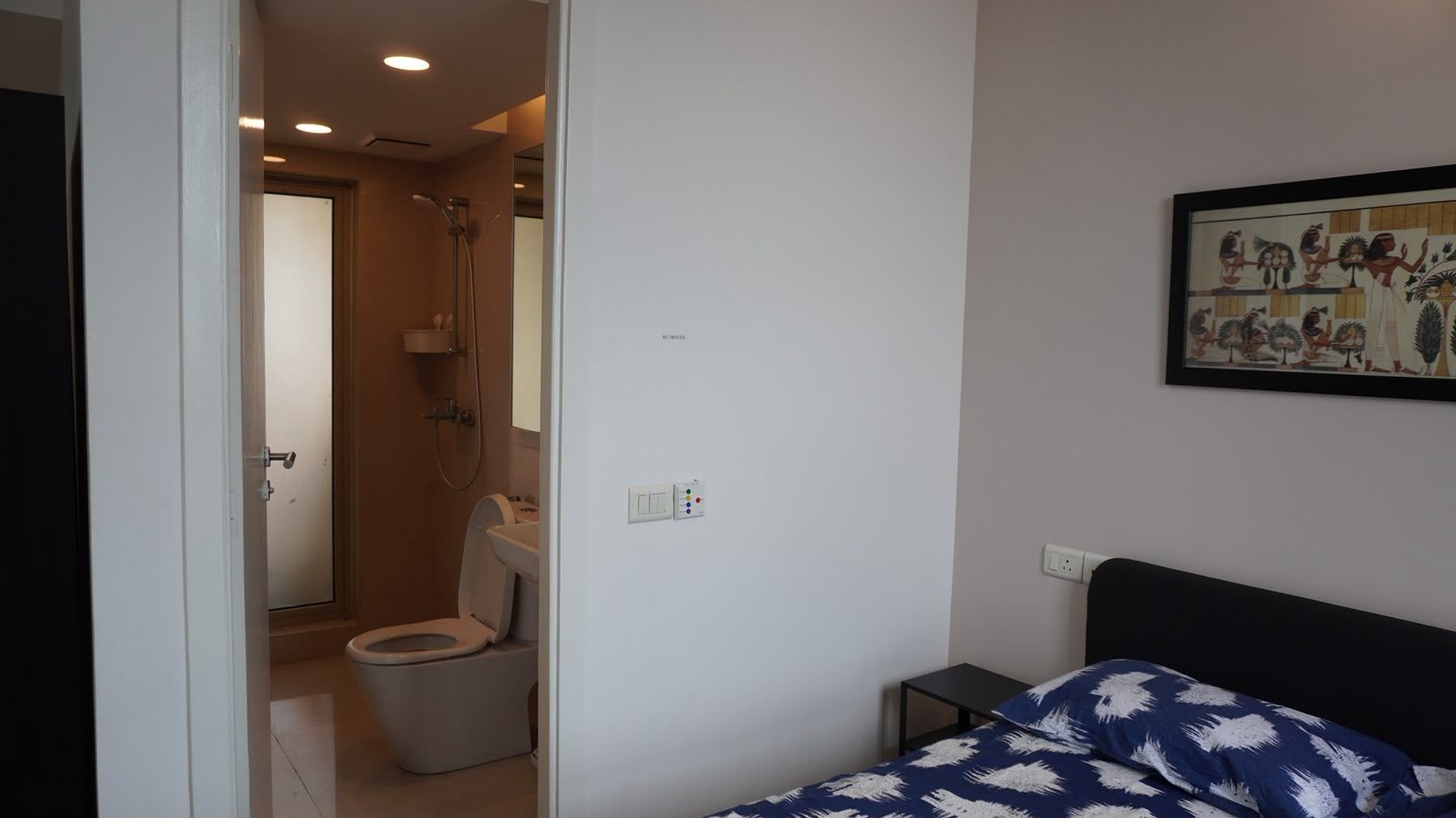 Ein Schlafzimmer mit einem Bett, einem kleinen Nachttisch, einem Wandbild und einem Badezimmer mit Toilette und Dusche.