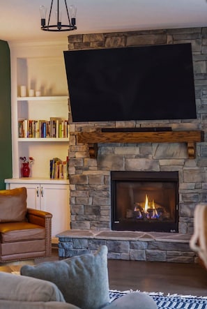 Ein Wohnzimmer mit Kamin aus Stein, einem flachbildfernseher darüber angebracht, einem Ledersessel und einem Bücherregal voller Bücher.