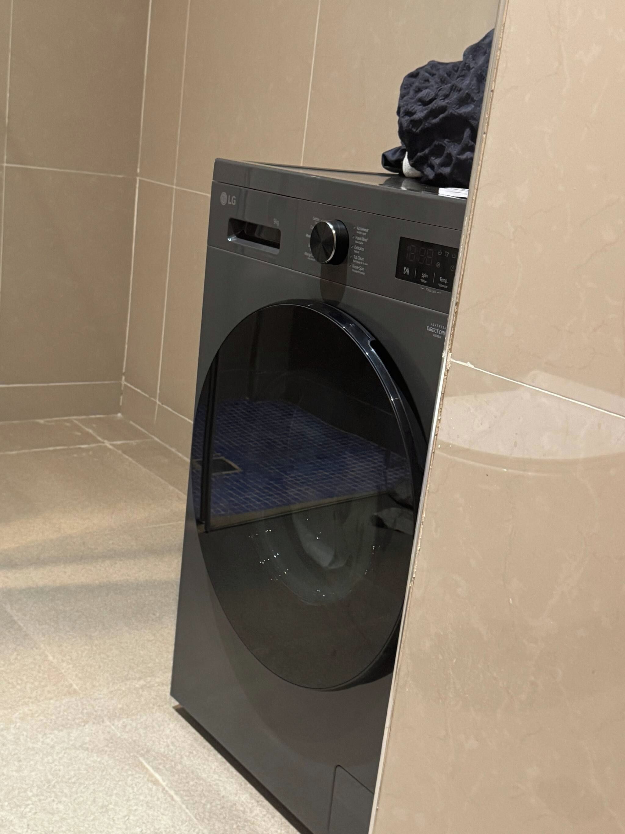 Une machine à laver LG à chargement frontal, installée dans une salle de bain aux carreaux et au sol carrelés, le tout dans des tons beige.