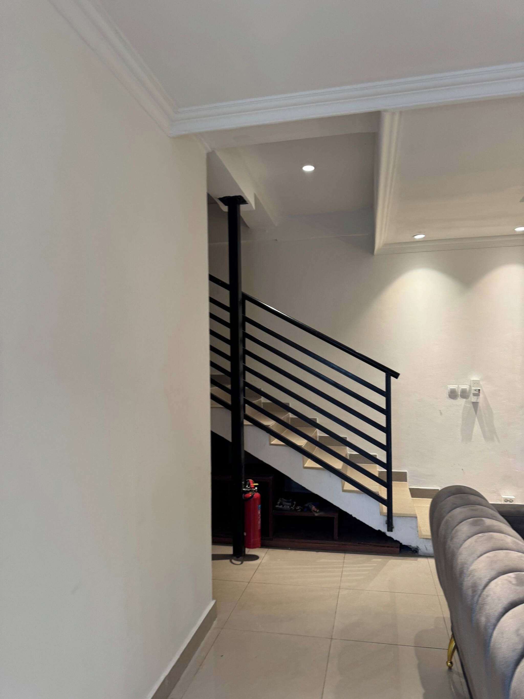 A staircase with a black railing leading to an upper floor, a fire extinguisher on the floor, and a grey sofa in the foreground.