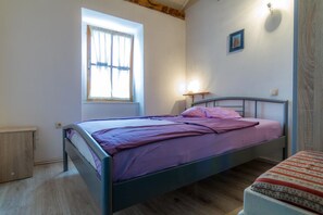 Uma cama de solteiro com cabeceira, janela com cortinas, abajur e mesa de cabeceira pequena.