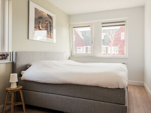 Ein Schlafzimmer mit einem Einzelbett, einem kleinen Hocker, einem gerahmten Bild an der Wand und einem Fenster mit Blick auf Gebäude.