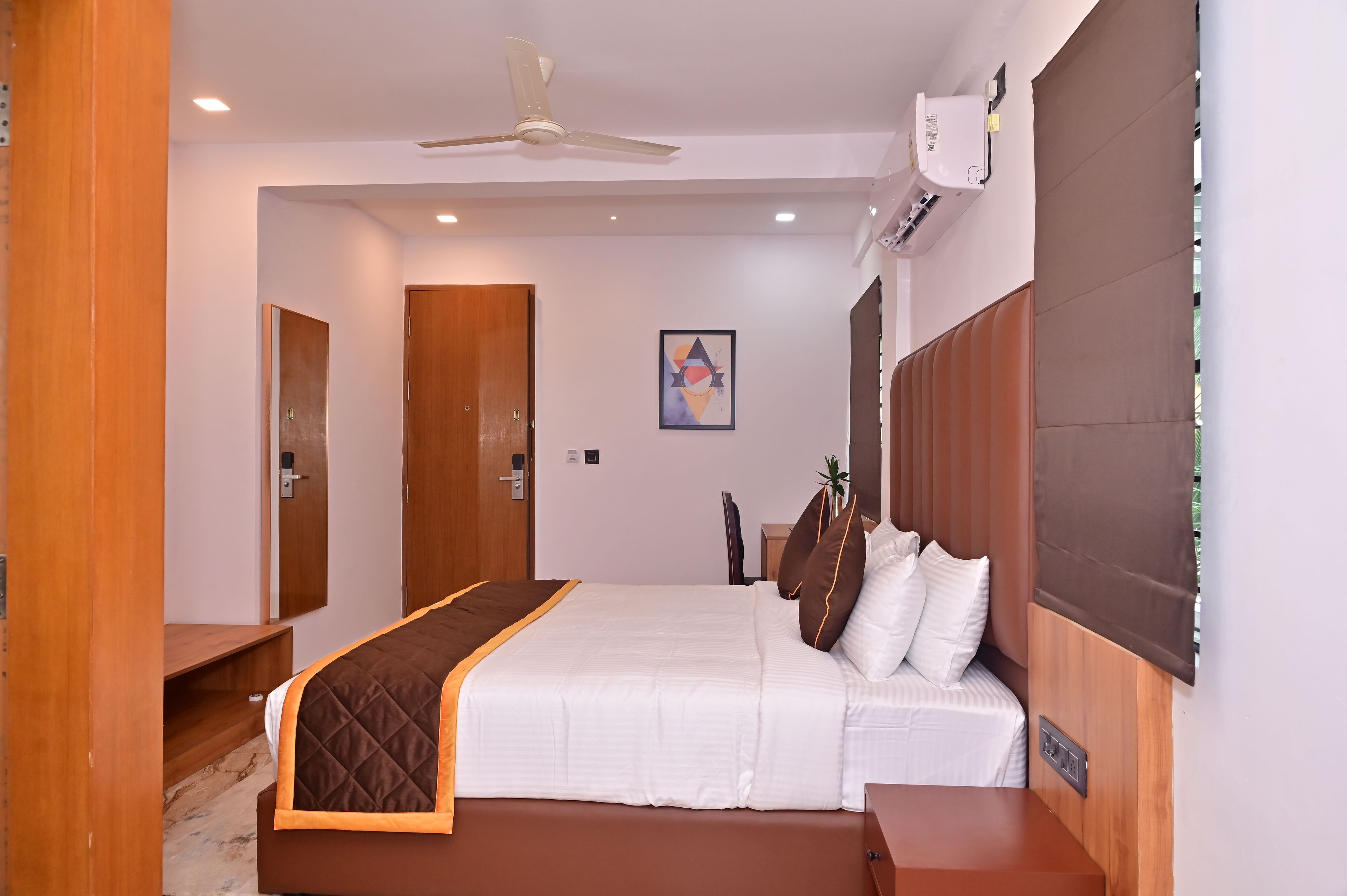 A hotel room with a bed, bedside table, wardrobe, ceiling fan, air conditioning unit, and a painting on the wall.