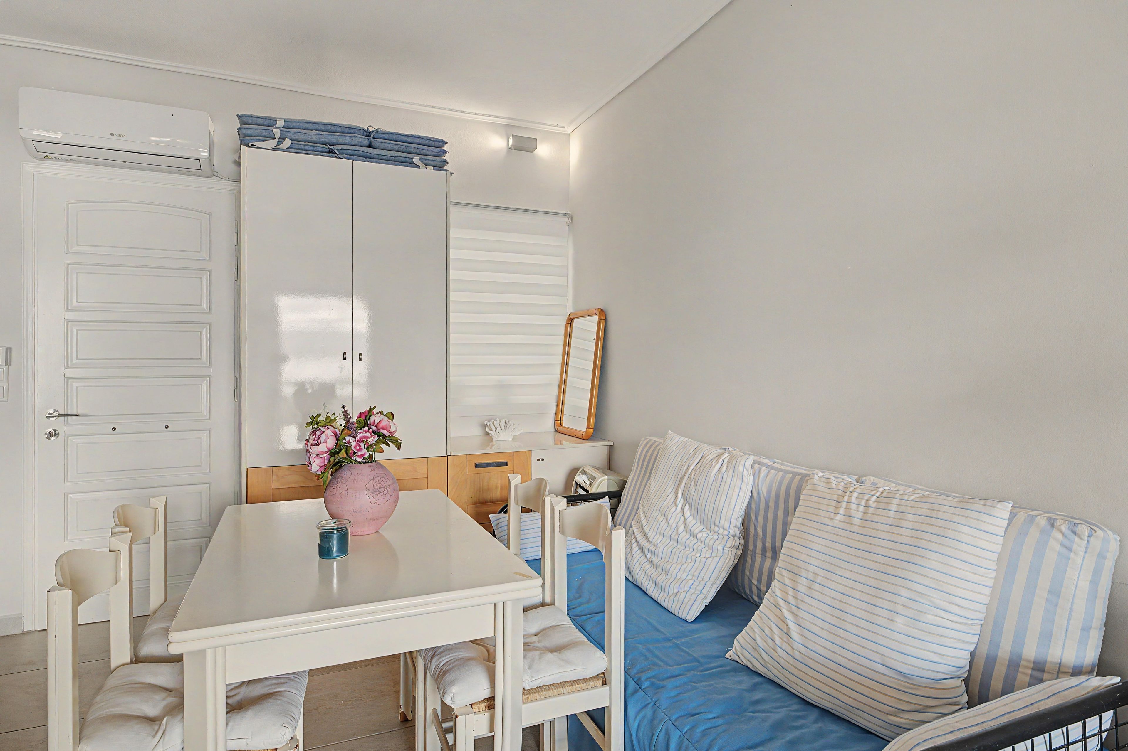 Een kamer met een witte tafel, stoelen, een bank met gestreepte kussens, een roze vaas met bloemen en een witte garderobe met spiegel.