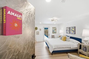 A hotel room with a bed, bedside table, lamp, and artwork on the wall. - Court Yard Suite | Pool | Downtown ILM (Wilmington)