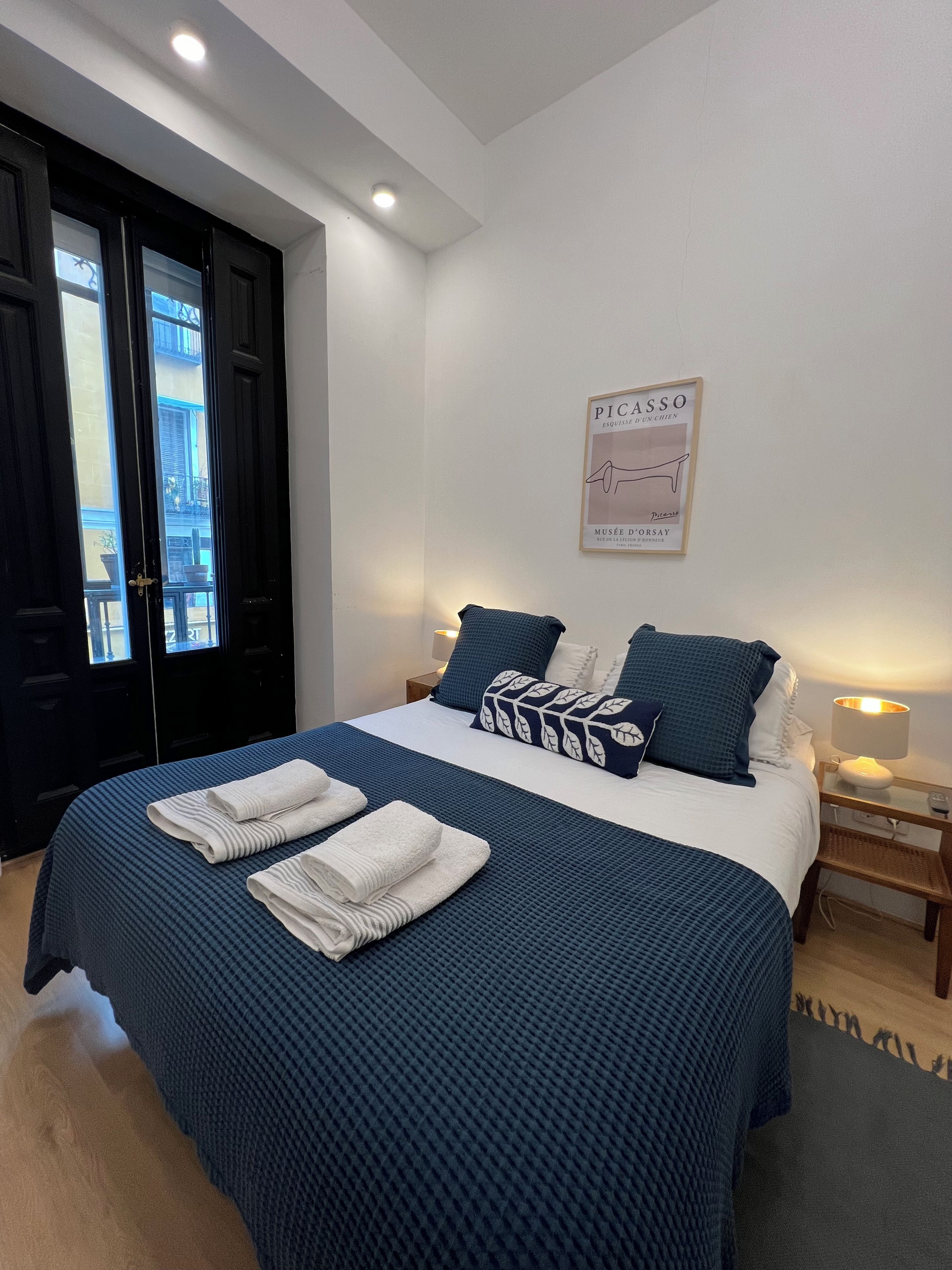 Una camera d'hotel con un letto, un comodino, una lampada a parete, un quadro incorniciato e una porta che si affaccia all'esterno.