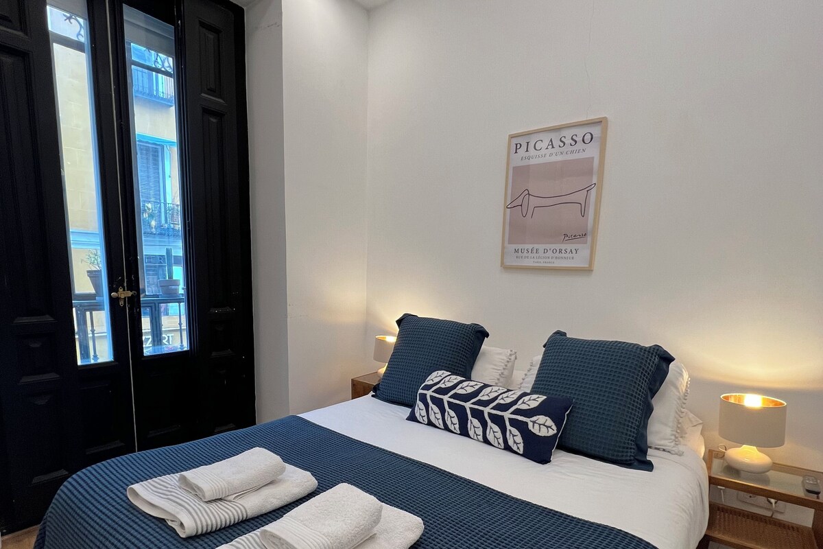 A hotel room with a bed, a nightstand, a wall-mounted lamp, a framed picture, and a door with a view of the outside.