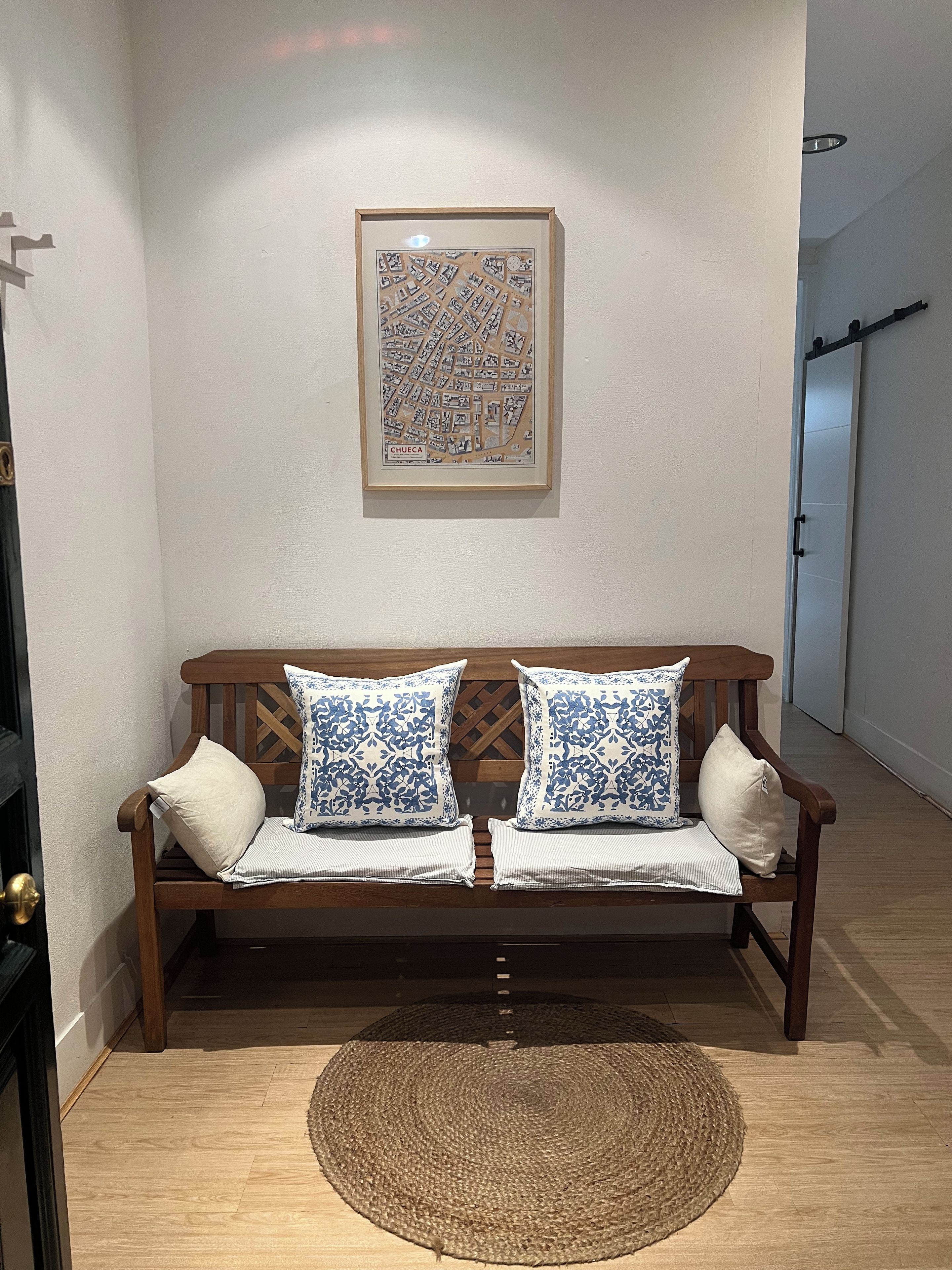 Un banc en bois avec des coussins à motifs adossé à un mur, une carte encadrée au-dessus, et un tapis tissé sur le sol.