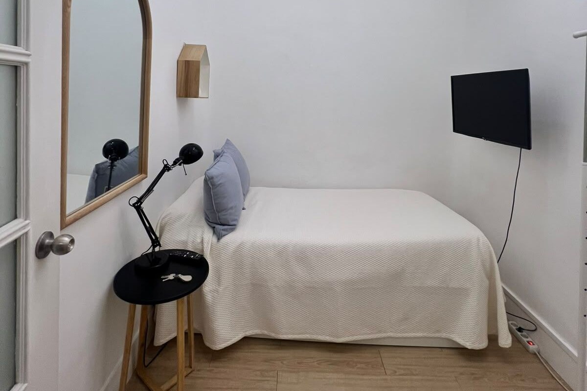 A small, neatly arranged bedroom with a single bed, a desk with a chair, a mirror, and a flat-screen TV mounted on the wall.