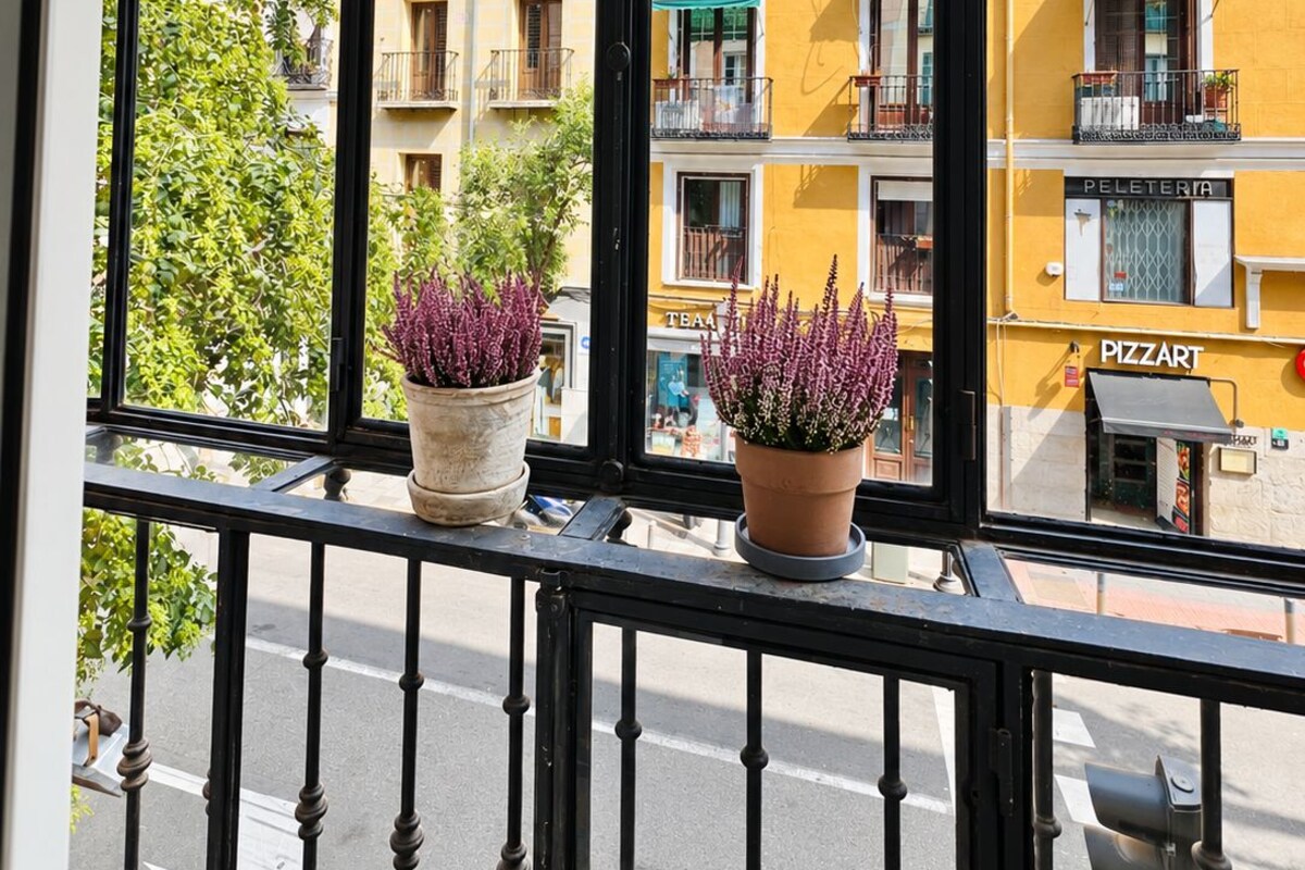 A balcony with a view of yellow buildings, a street, and two potted plants.