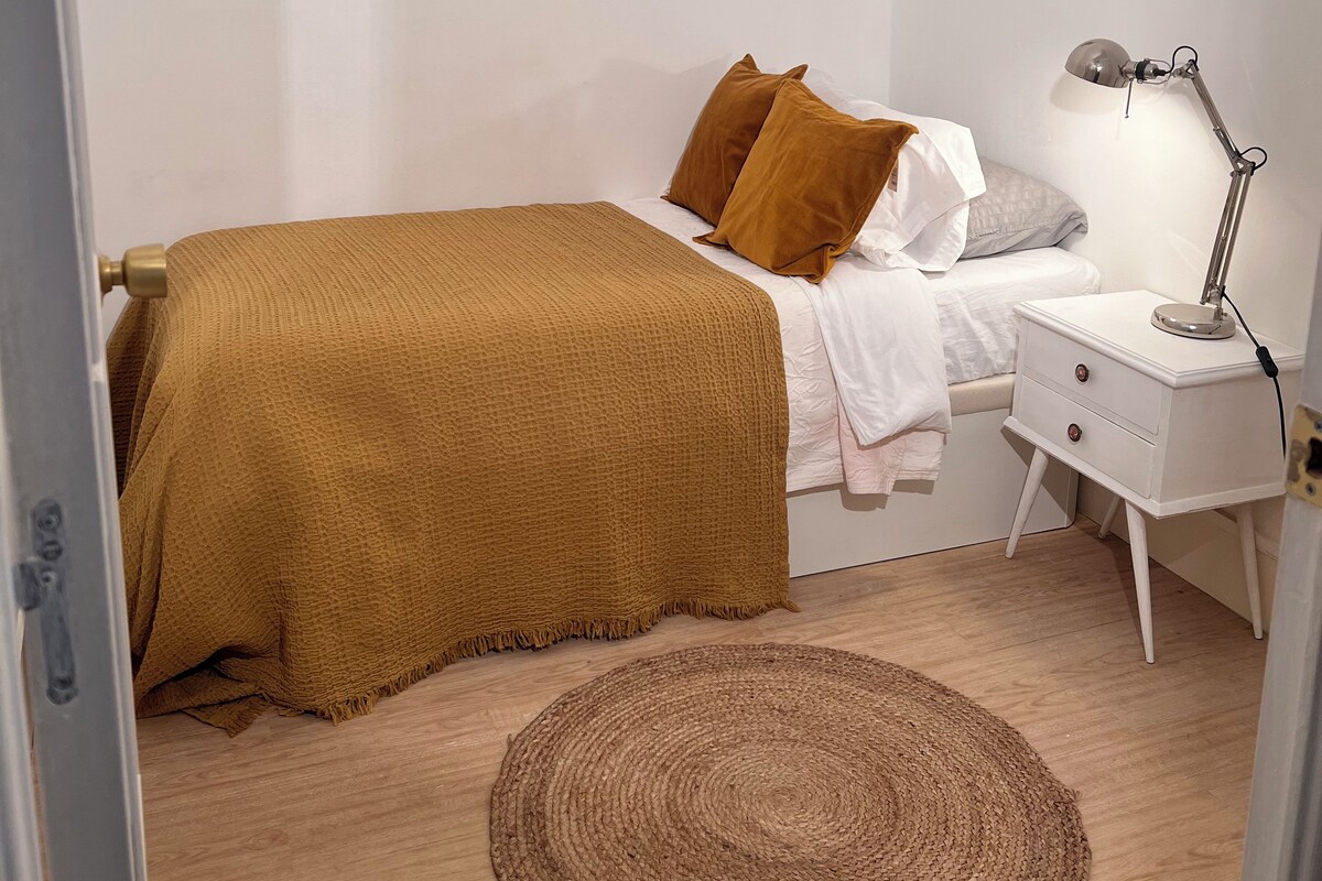 A bedroom with a bed, bedside table, lamp, and a small wooden shelf.