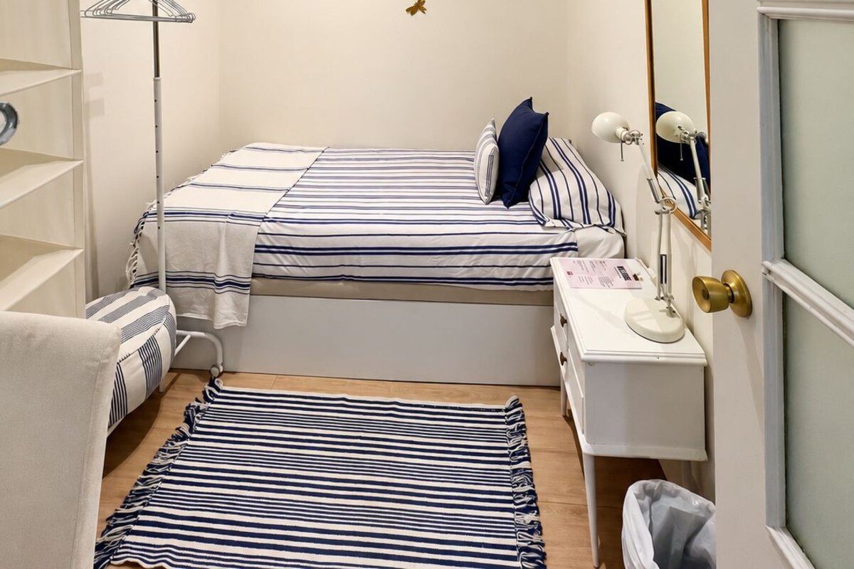 A small, well-lit bedroom with a bed, bedside table, lamp, and a mirror.
