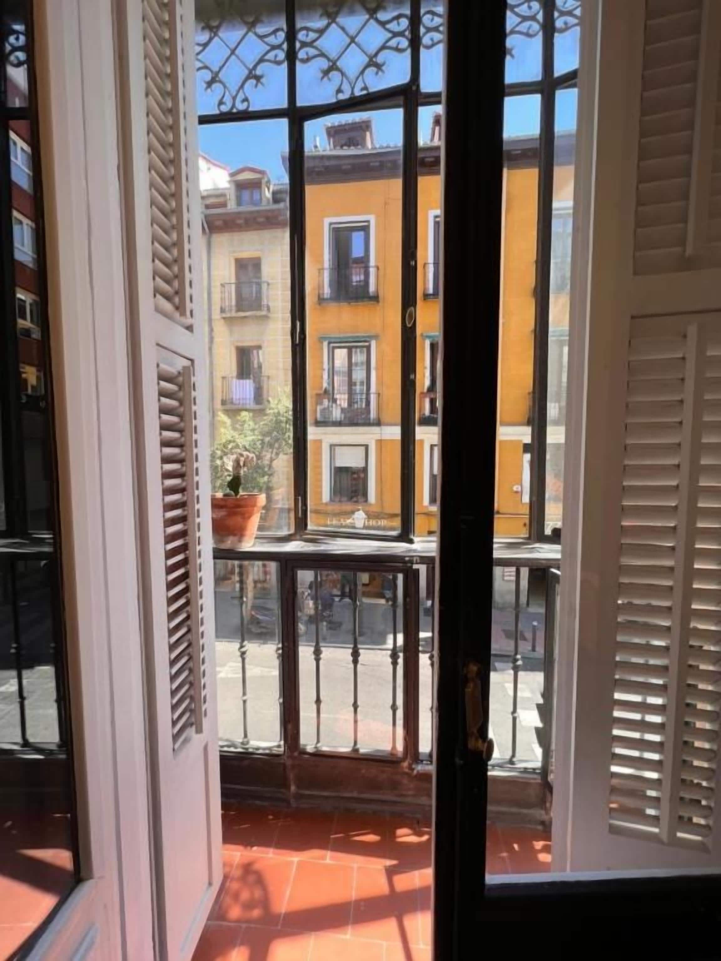Blick durch ein Fenster mit einem Metallgitter, das auf einen Balkon mit Fliesenboden und einer Topfpflanze führt.