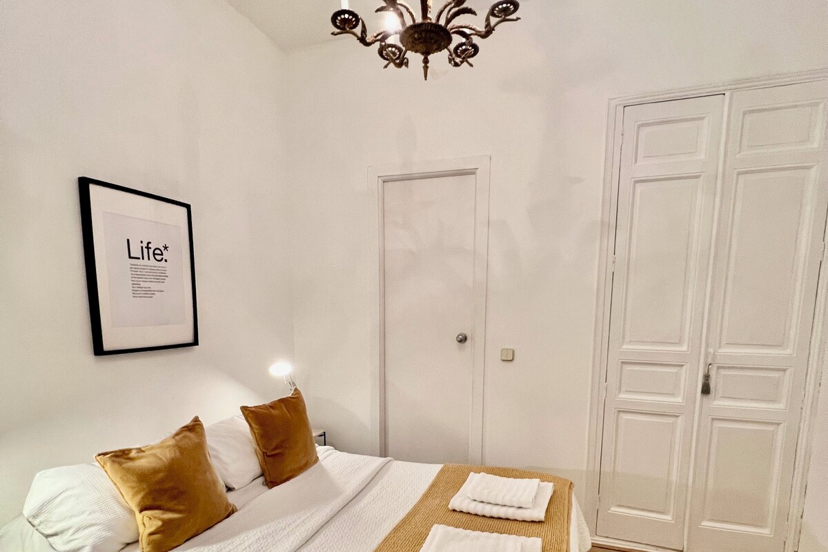 A neatly made bed with white linens and a brown throw blanket, a framed picture on the wall, and a closed door in the background.