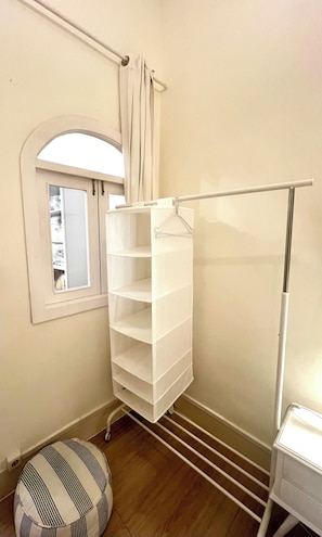 A room with a white storage unit, a window with curtains, a striped ottoman, and a white radiator.