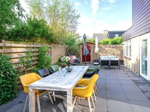 A patio with a wooden table and yellow chairs, surrounded by greenery and a brick wall. - Holiday Home With 2 Bedrooms (De Cocksdorp)