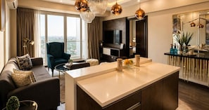 Una sala moderna con un sofá, una mesa de centro y un televisor. Cuenta con un ventanal con cortinas, un candelabro y una zona de cocina con mostrador y armarios.