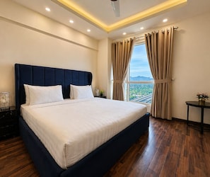 Habitación de hotel con una cama grande, una mesita de noche, una pequeña mesa con un jarrón y una ventana con cortinas.