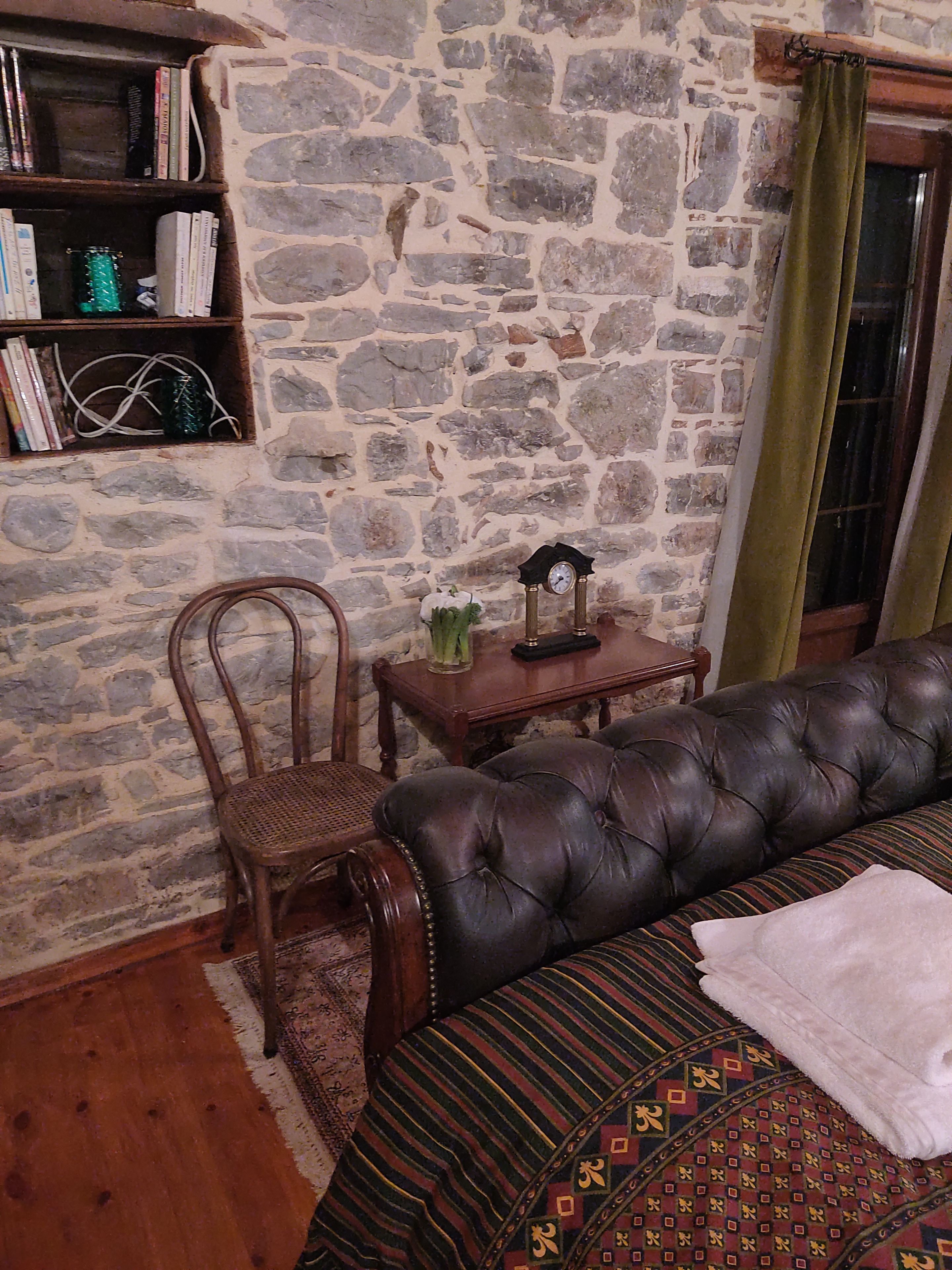 A room with a stone wall, a leather sofa, a wooden chair, a small table with a clock, and a bookshelf.