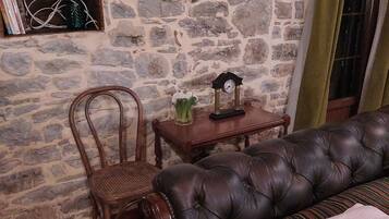 A room with a stone wall, a leather sofa, a wooden chair, a small table with a clock, and a bookshelf.
