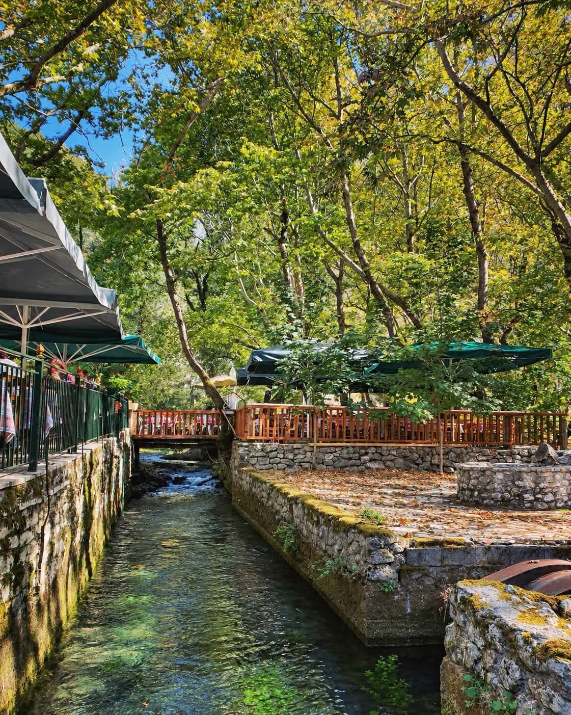 A riverside walkway with stone walls, a wooden bridge, and a restaurant with outdoor seating.