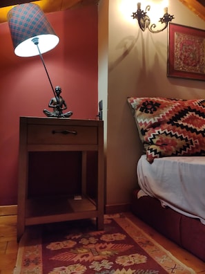 A room with a bed, a nightstand with a lamp, a wall-mounted light fixture, and a patterned rug.