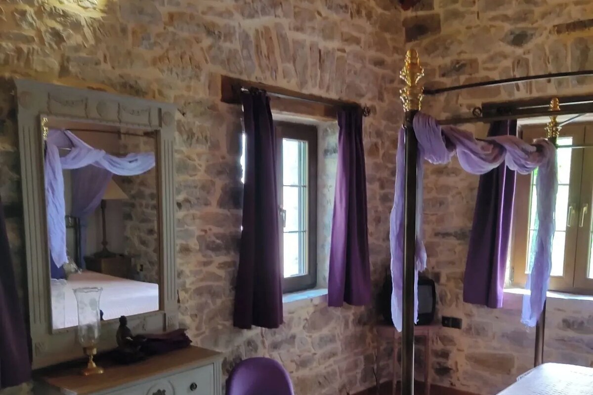 Een kamer met stenen muren, een bed, een dressoir, een stoel en een raam met gordijnen.