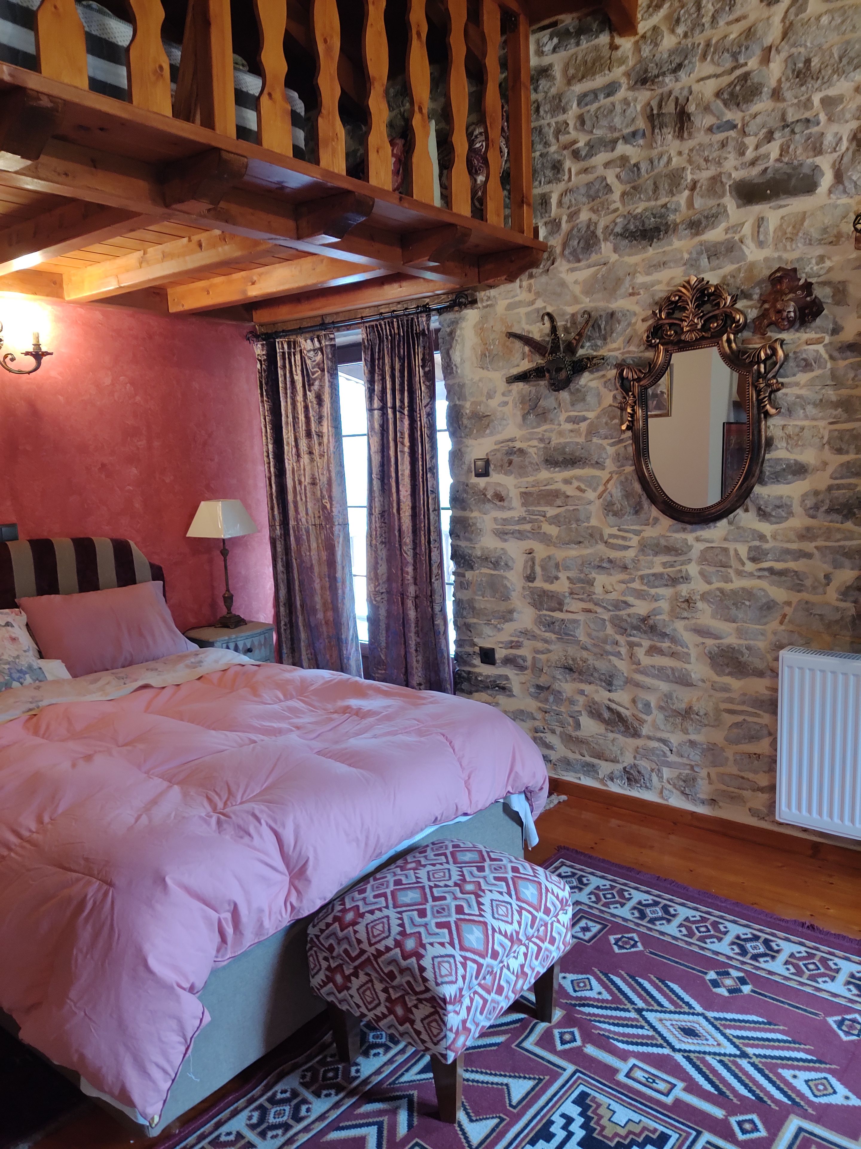 A bedroom with a bed, a nightstand, a lamp, a patterned rug, a wooden bench, a stone wall, and a mirror.