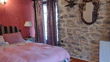 A bedroom with a bed, a nightstand, a lamp, a patterned rug, a wooden bench, a stone wall, and a mirror.
