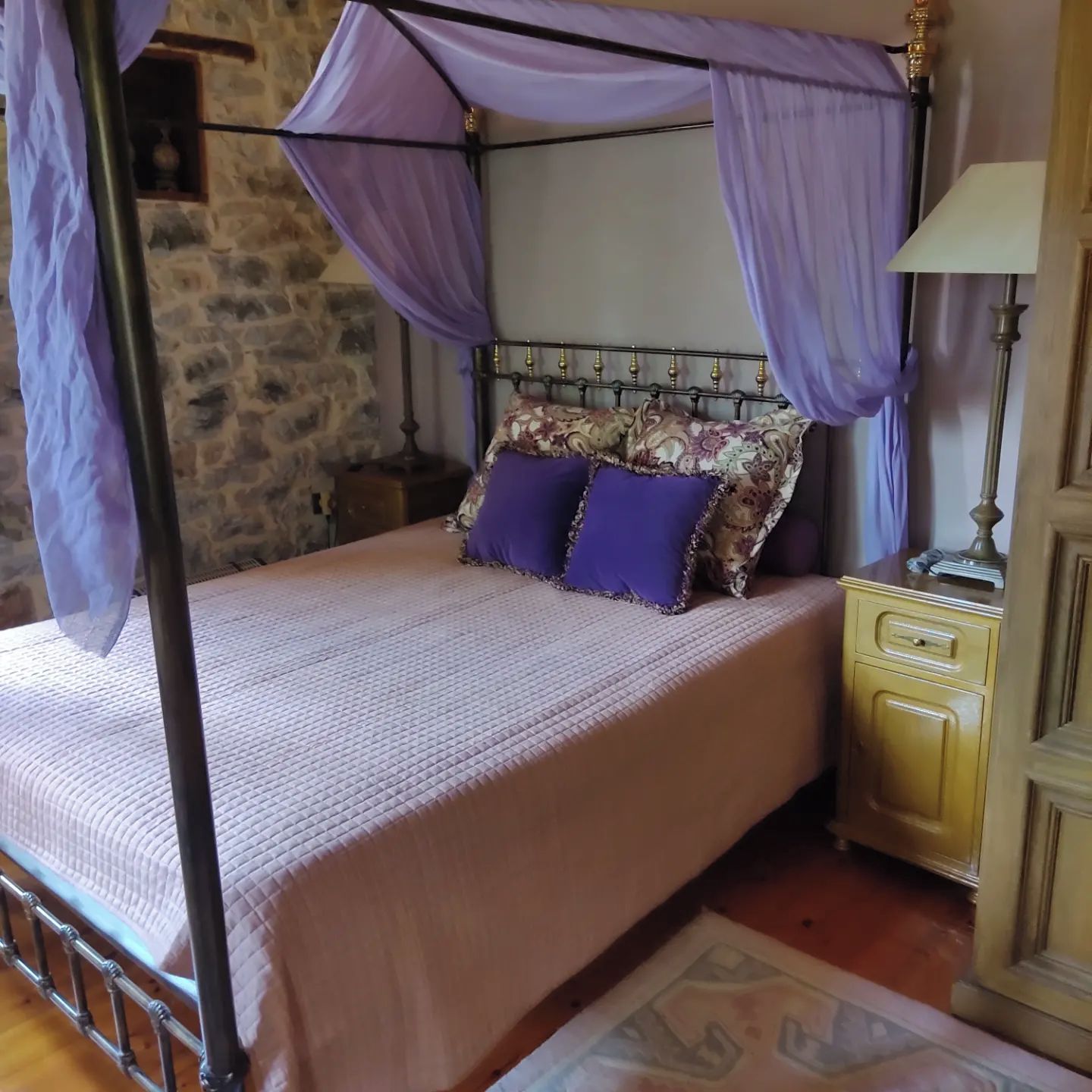 Una cama con dosel, una mesita de noche, una lámpara y una pared de piedra.