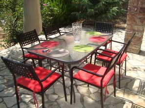 A glass-top outdoor table with red cushioned chairs, surrounded by stone flooring and greenery.