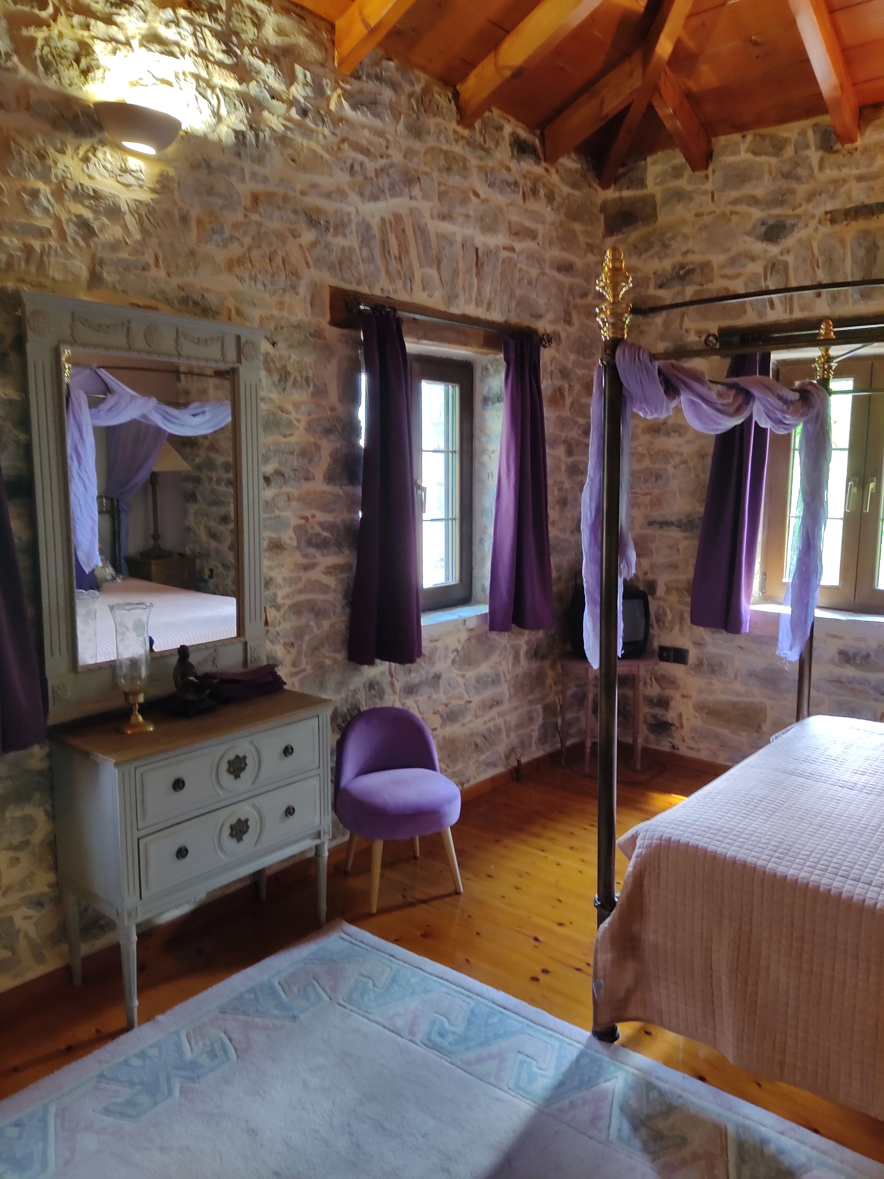 Un dormitorio con paredes de piedra, techo de madera, cama con cabecera, mesita de noche, cómoda, silla y ventana con cortinas.