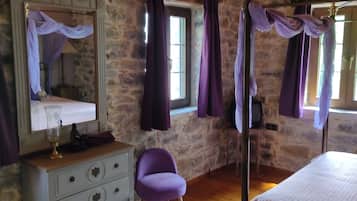 Un dormitorio con paredes de piedra, techo de madera, cama con cabecera, mesita de noche, cómoda, silla y ventana con cortinas.