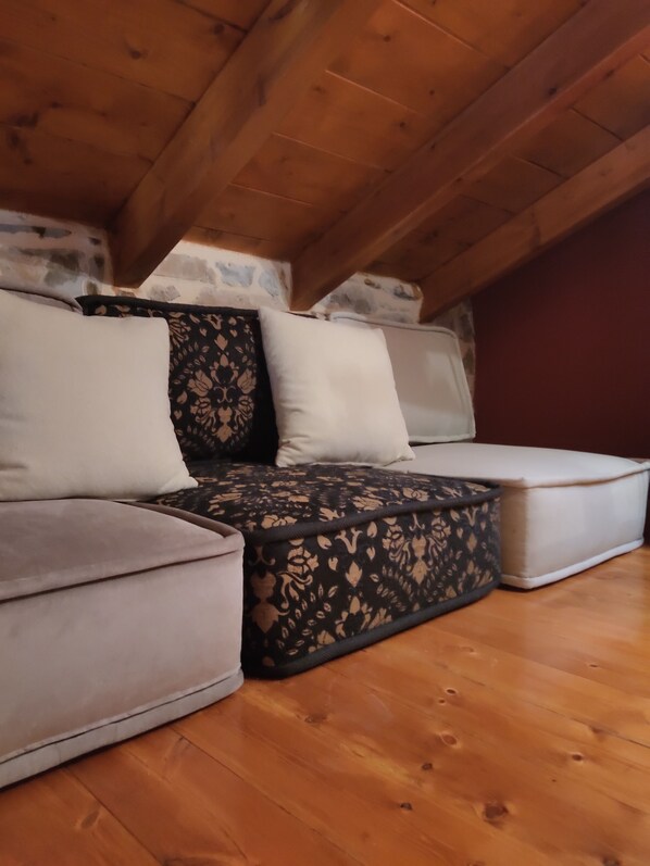 A wooden ceiling with a sloped roof, a sofa with patterned cushions, and a white ottoman.