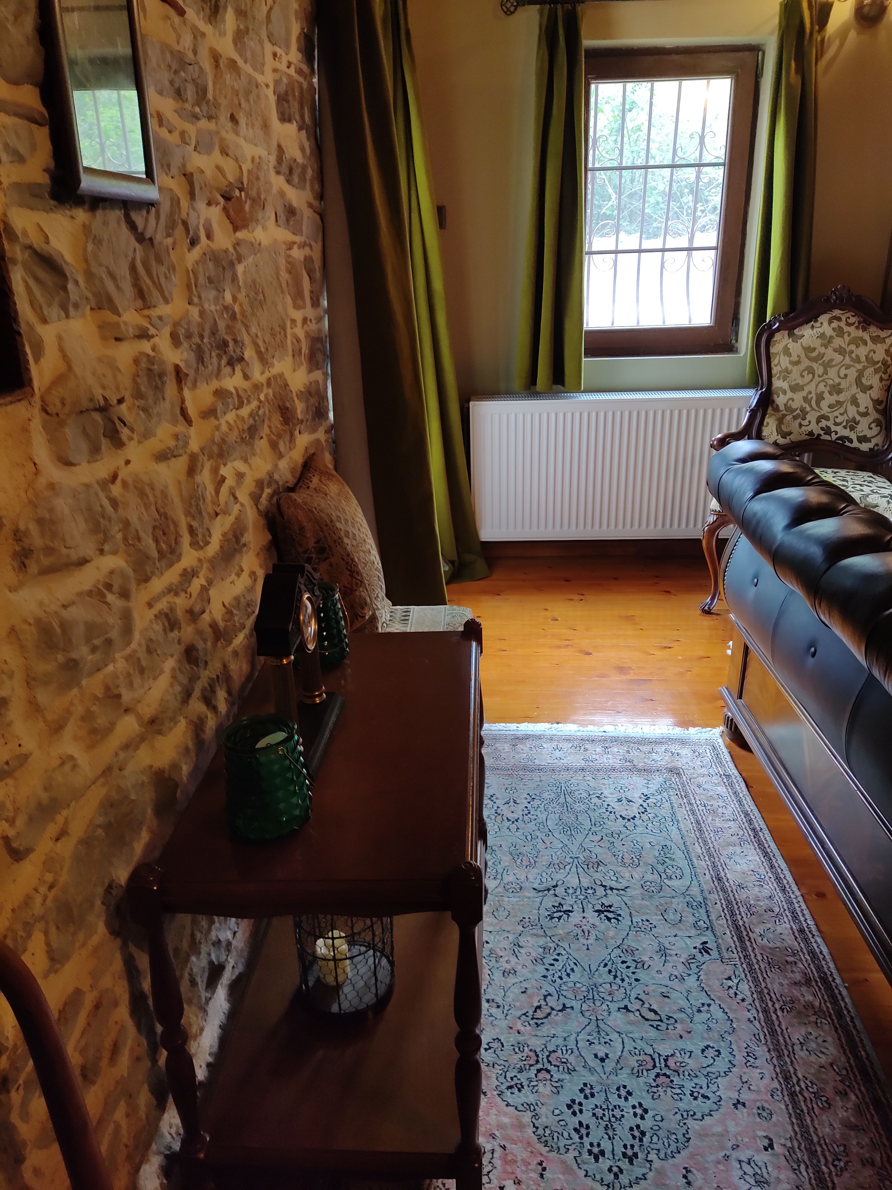 Una habitación acogedora con pared de piedra, una mesa de madera con vela, alfombra estampada, sofá de cuero y una ventana con cortinas.